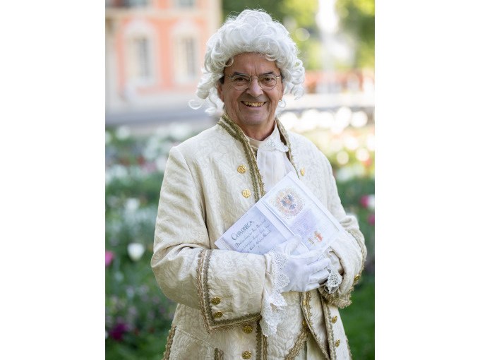 Ein Mann in barocker Kleidung und weißer Perücke steht lächelnd in einem Garten und hält ein Buch. Im Hintergrund sind Blumen und ein Gebäude zu sehen., © Touristik und Marketing GmbH Ein Mann in barocker Kleidung und weißer Perücke steht lächelnd in einem Garten und hält ein Buch. Im Hintergrund sind Blumen und ein Gebäude zu sehen., © Touristik und Marketing GmbH