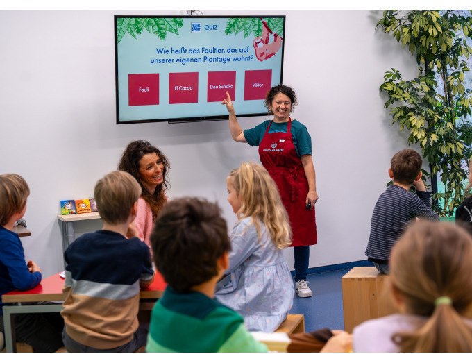 Kinder sitzen in einem Raum und machen ein Quiz über Faultiere. Eine Frau in roter Schürze steht vorne und zeigt auf den Bildschirm., © Alfred Ritter GmbH & Co. KG | Waldenbuch Kinder sitzen in einem Raum und machen ein Quiz über Faultiere. Eine Frau in roter Schürze steht vorne und zeigt auf den Bildschirm., © Alfred Ritter GmbH & Co. KG | Waldenbuch