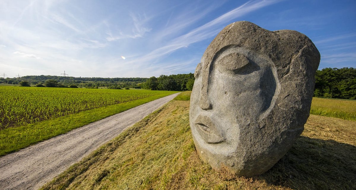 Steinskulptur eines Gesichts am Oeffinger Rundweg, neben einem Feldweg und einem Feld unter blauem Himmel., © Remstal Tourismus e.V. Steinskulptur eines Gesichts am Oeffinger Rundweg, neben einem Feldweg und einem Feld unter blauem Himmel., © Remstal Tourismus e.V.