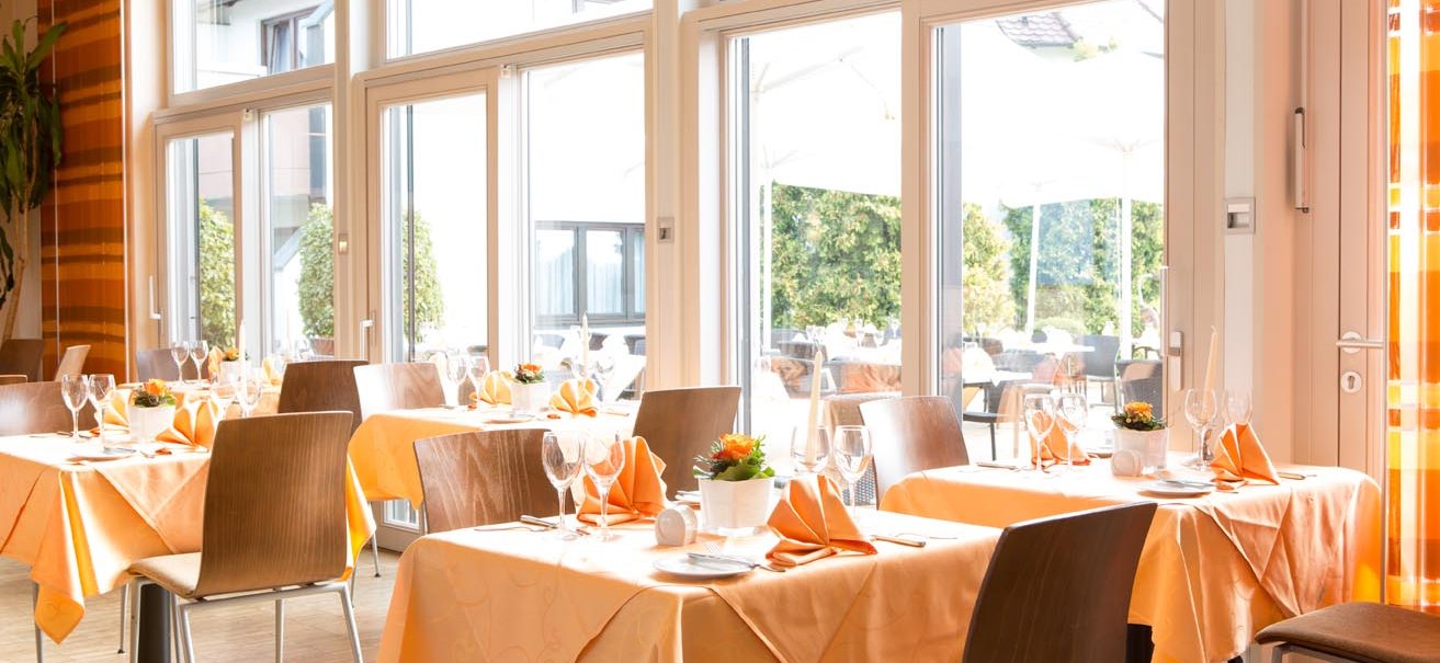 Helles Restaurant mit großen Fenstern, orange Tischdecken und elegant gedeckten Tischen. Außenbereich mit Sonnenschirmen sichtbar., © ARAMIS Hotel & Freizeit GmbH