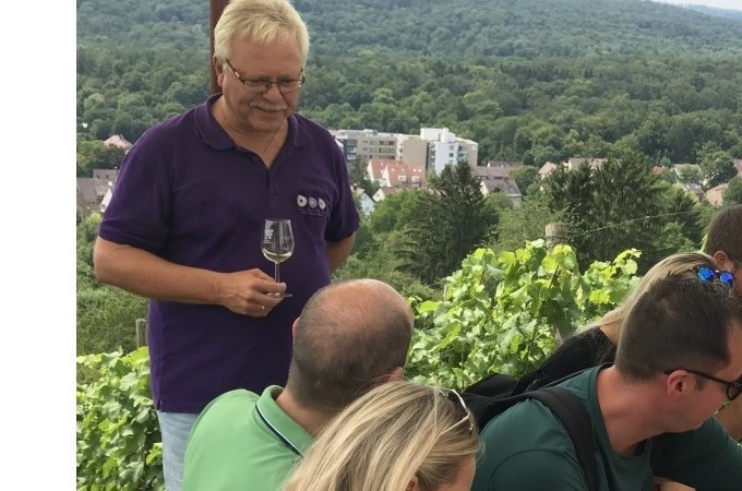 Ein Mann in lila Hemd hält ein Weinglas und spricht mit einer Gruppe von Menschen in einem Weinberg. Im Hintergrund sind Bäume und Gebäude zu sehen., © Weingenuss Stuttgart