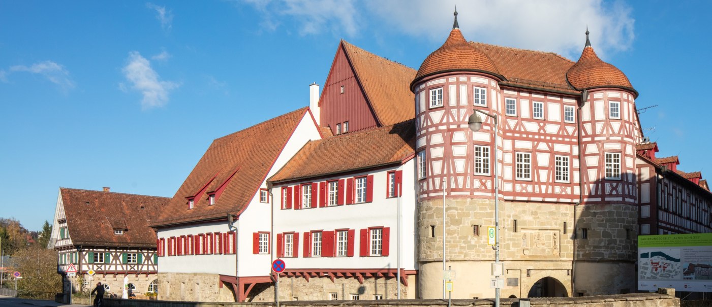 altes Schloss - rechts das Torhaus mit Sockelgeschoss aus hellem Sandstein und aufgesetztem roten Fachwerk mit weißen Gefachen, flankiert von zwei Rundtürmen. In der Bildmitte der Westflügel mit zwei aneinandergebauten, weiß verputzten Gebäudeteilen mit roten Fensterläden, überragt vom Dach und rotbraun gestrichener Giebelseite des Nordflügels. Alle Gebäudeteile eingedekct mit roten Ziegeln. Links das Fachwerkgebäude der ehemaligen Bentheimschen Forstverwaltung mit hellem Sandsteinsockel und grünen Fensterläden., © Stadt Gaildorf