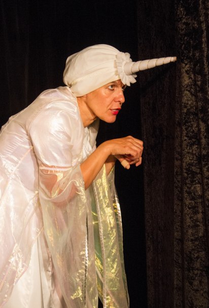 Eine Person in einem wei&szlig;en, schimmernden Kost&uuml;m mit einem Einhorn-Hut spielt auf einer Theaterb&uuml;hne. Der Hintergrund ist dunkel gehalten., &copy; Galli Theater Backnang