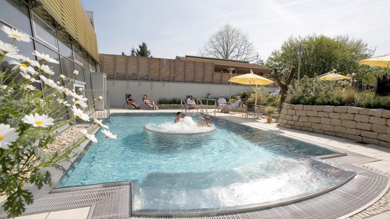 Außenbereich der Barbarossa-Thermen mit Pool, sprudelndem Wasser und Liegestühlen. Menschen entspannen unter Sonnenschirmen. Blumen und Bäume im Hintergrund., © Barbarossa-Thermen Göppingen