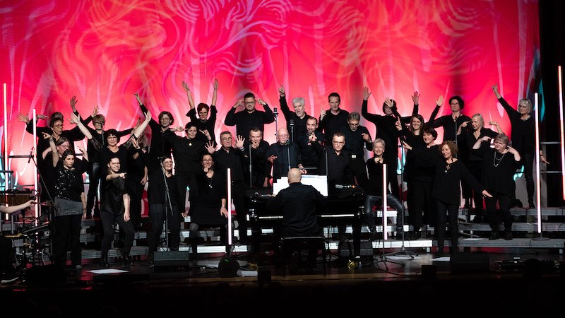 Ein Chor in schwarzer Kleidung singt auf einer Bühne mit rotem Hintergrund. Ein Dirigent spielt Klavier. Die Beleuchtung ist stimmungsvoll., © Gesangverein Ernsbach e.V.