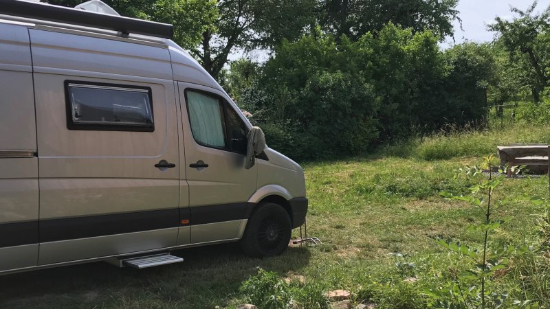 Ein silbernes Wohnmobil steht auf einem grünen Stellplatz. Im Hintergrund sind Bäume und eine rustikale Bank zu sehen., © Weingut Idler Ein silbernes Wohnmobil steht auf einem grünen Stellplatz. Im Hintergrund sind Bäume und eine rustikale Bank zu sehen., © Weingut Idler