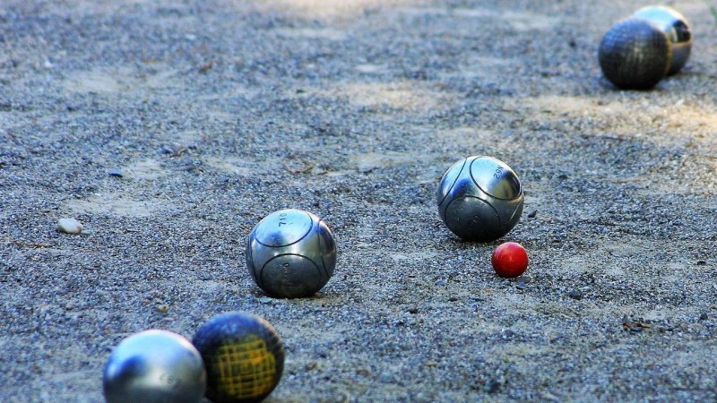 Metallene Boule-Kugeln liegen auf einem Sandplatz um eine kleine rote Zielkugel verteilt. Das Bild zeigt eine typische Spielsituation beim Boule., &copy; boule-3058654_1280_JuergenSieber_Pixabay