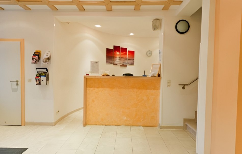 Ein Empfangsbereich mit einem Tresen, Wandbild, Broschürenhalter und einer Uhr an der Wand. Helle, freundliche Atmosphäre., © Hotel Neuwirtshaus