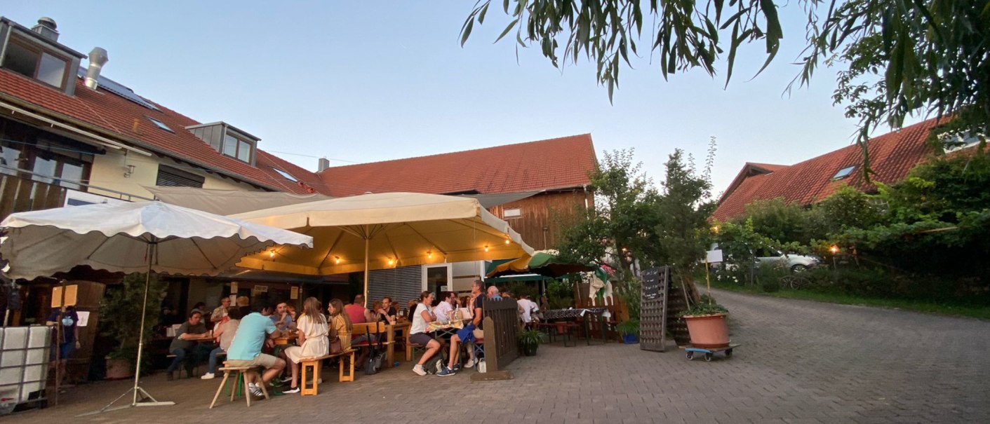Menschen sitzen unter gro&szlig;en Sonnenschirmen in einem gem&uuml;tlichen Hof mit roten D&auml;chern und B&auml;umen., &copy; Weinhof Zai&szlig;