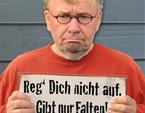 Mann mit Brille und rotem Pullover hält ein Schild mit der Aufschrift: 'Reg' Dich nicht auf. Gibt nur Falten!' vor einem blauen Hintergrund., © Scala Kultur Live gGmbH Mann mit Brille und rotem Pullover hält ein Schild mit der Aufschrift: 'Reg' Dich nicht auf. Gibt nur Falten!' vor einem blauen Hintergrund., © Scala Kultur Live gGmbH