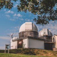 Ein Observatorium mit zwei Kuppeln steht auf einem Hügel unter blauem Himmel. Bäume rahmen das Bild ein., © Stadt Welzheim