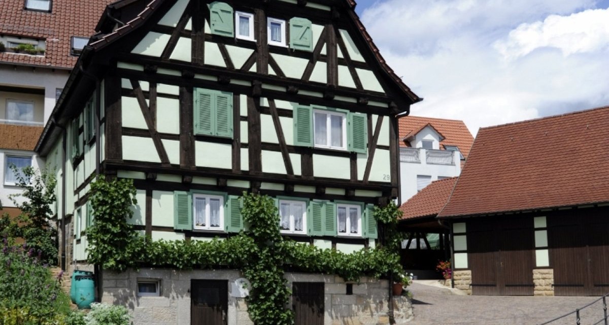 Fachwerkhaus mit gr&uuml;nem Anstrich und blauen Fensterl&auml;den, umgeben von einem gepflasterten Hof und bl&uuml;henden Pflanzen, unter blauem Himmel., &copy; Natur.Nah. Sch&ouml;nbuch & Heckeng&auml;u