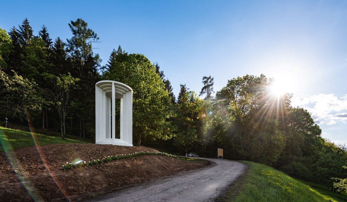 Weiße Säulenstruktur auf einem Hügel neben einem Weg, umgeben von Bäumen und Sonnenschein., © Authentic Studios Weiße Säulenstruktur auf einem Hügel neben einem Weg, umgeben von Bäumen und Sonnenschein., © Authentic Studios