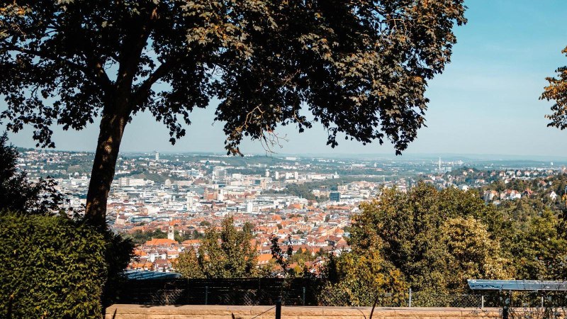 Panoramablick auf eine Stadt mit Bäumen im Vordergrund, die den Blick einrahmen. Eine Person sitzt auf einer Bank und genießt die Aussicht., © Stuttgart-Marketing GmbH, Sarah Schmid