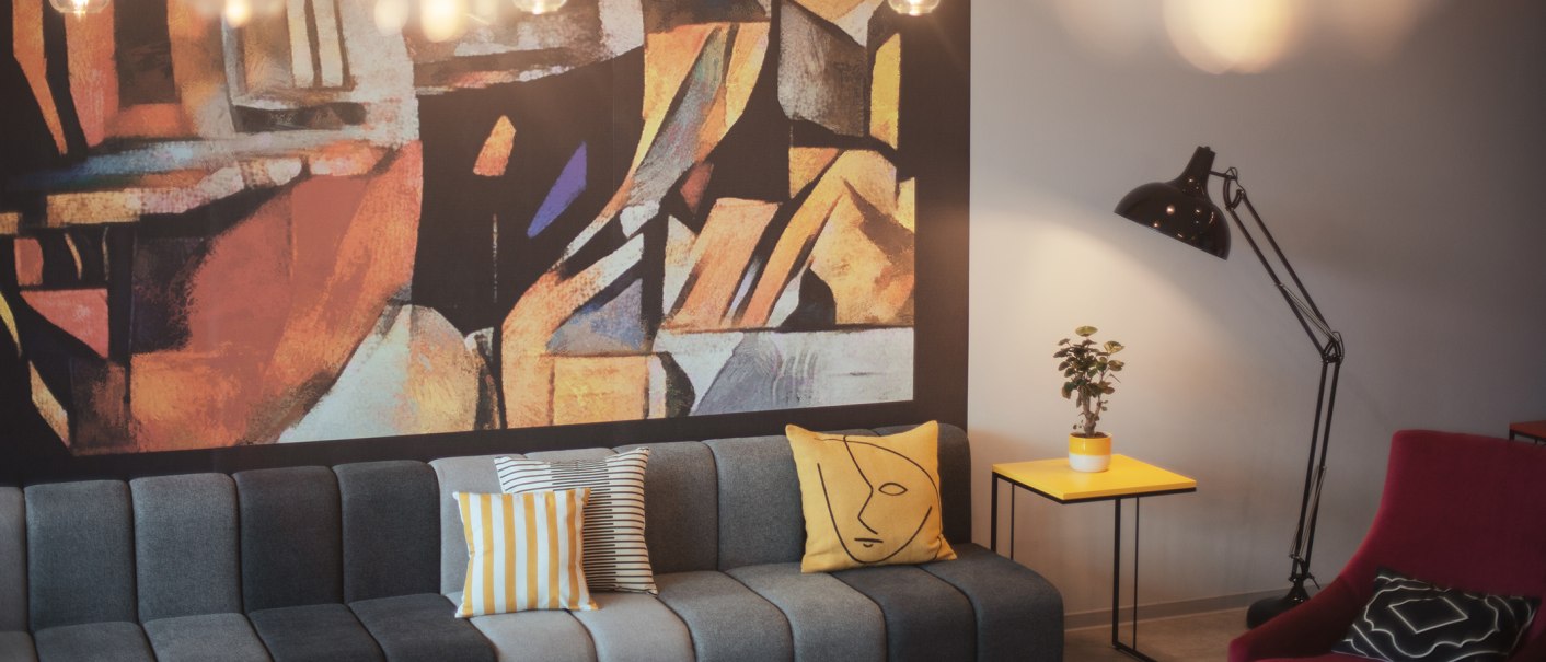 Moderne Lobby mit grauem Sofa, bunten Kissen, abstraktem Wandbild, gelbem Beistelltisch, Pflanze und Stehlampe., © the niu Form - Novum Hospitality