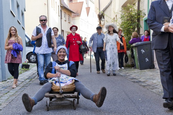 Frau auf kleinem Wagen in schmaler Gasse, umgeben von fröhlichen Menschen. Historische Kleidung und entspannte Atmosphäre., © Stadt Besigheim