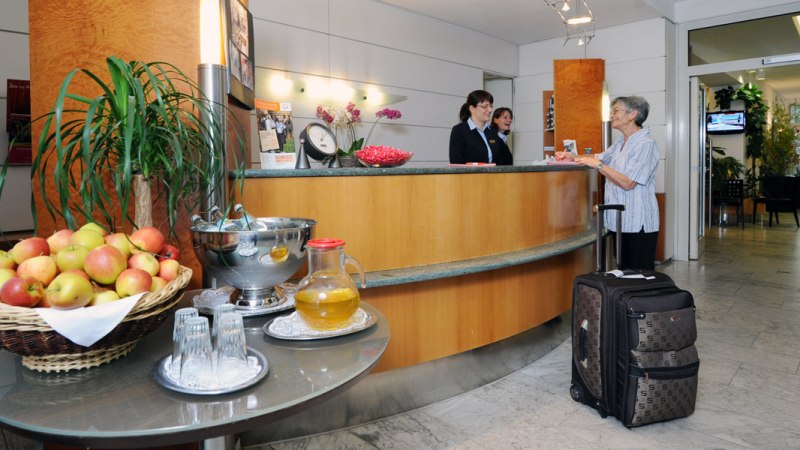 Eine Hotelrezeption mit zwei Mitarbeiterinnen, die einer &auml;lteren Frau mit Koffer helfen. Ein Tisch mit &Auml;pfeln und Getr&auml;nken steht im Vordergrund., &copy; Hotel Unger