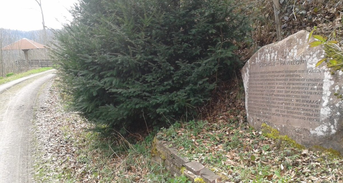 Ein Gedenkstein mit Inschrift steht am Rand eines Waldwegs. Ein gro&szlig;er Busch w&auml;chst daneben. Im Hintergrund ist ein Pavillon zu sehen., &copy; Natur.Nah. Sch&ouml;nbuch & Heckeng&auml;u