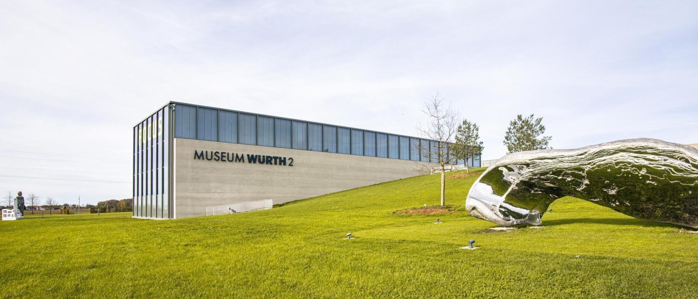 Das Museum Würth 2 in Künzelsau zeigt moderne Architektur mit einer großen, glänzenden Skulptur im Vordergrund auf einer grünen Wiese., © SMG, Sarah Schmid Das Museum Würth 2 in Künzelsau zeigt moderne Architektur mit einer großen, glänzenden Skulptur im Vordergrund auf einer grünen Wiese., © SMG, Sarah Schmid