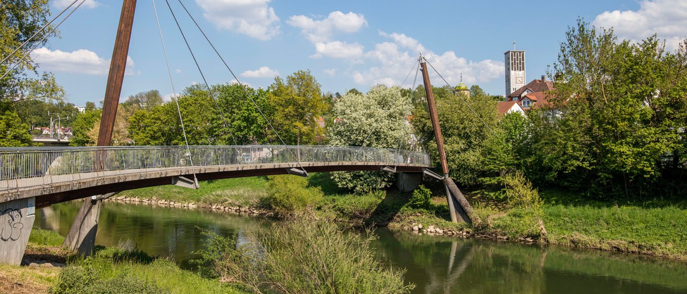 Eine moderne Fußgängerbrücke überspannt einen Fluss, umgeben von grüner Vegetation. Im Hintergrund ist ein Kirchturm zu sehen., © Stuttgart-Marketing GmbH Eine moderne Fußgängerbrücke überspannt einen Fluss, umgeben von grüner Vegetation. Im Hintergrund ist ein Kirchturm zu sehen., © Stuttgart-Marketing GmbH
