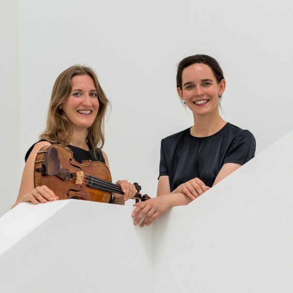 Zwei Frauen lächeln hinter einer weißen Wand, eine hält eine Geige. Sie tragen elegante, dunkle Kleidung., © Mauerwerk GmbH Zwei Frauen lächeln hinter einer weißen Wand, eine hält eine Geige. Sie tragen elegante, dunkle Kleidung., © Mauerwerk GmbH