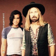 Zwei M&auml;nner stehen vor einem braunen Hintergrund. Einer tr&auml;gt einen Hut und hat langes blondes Haar, der andere hat dunkles, lockiges Haar., &copy; links im Bild