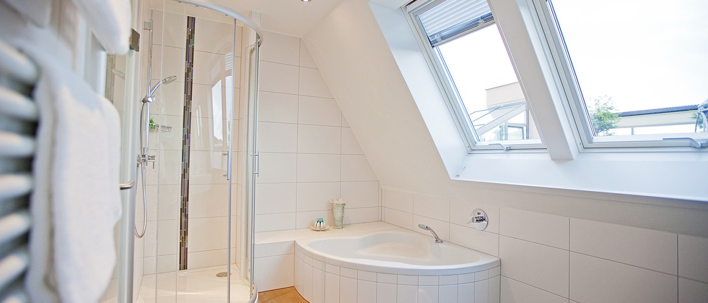 Helles Badezimmer mit Eckbadewanne, runder Duschkabine und großen Dachfenstern. Weiße Fliesen und Handtuchhalter an der Wand., © Hotel Krone Helles Badezimmer mit Eckbadewanne, runder Duschkabine und großen Dachfenstern. Weiße Fliesen und Handtuchhalter an der Wand., © Hotel Krone