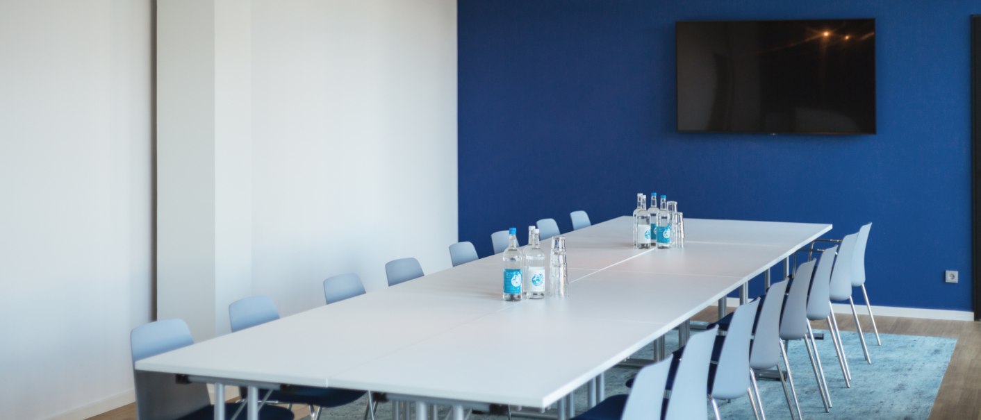 Ein moderner Meetingraum mit weißen Tischen und Stühlen, blauer Wand und Teppich. Auf dem Tisch stehen Wasserflaschen, an der Wand hängt ein Flachbildschirm., © Novum Management GmbH Ein moderner Meetingraum mit weißen Tischen und Stühlen, blauer Wand und Teppich. Auf dem Tisch stehen Wasserflaschen, an der Wand hängt ein Flachbildschirm., © Novum Management GmbH