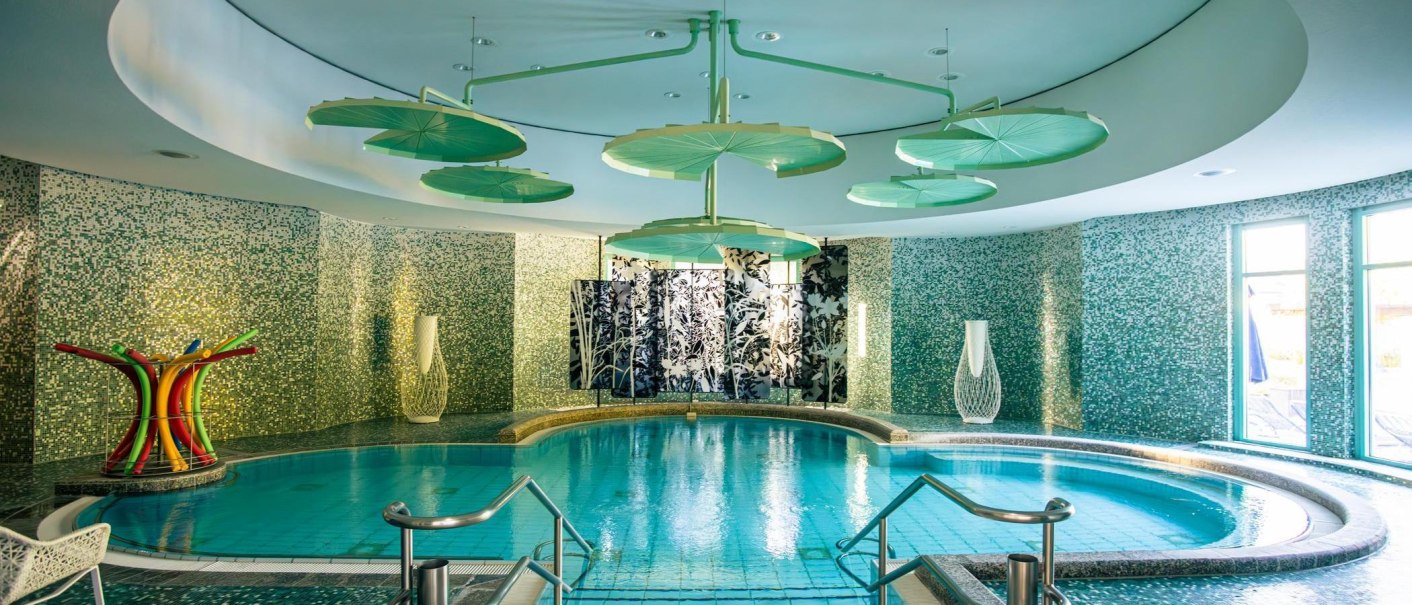 Ein eleganter Spa-Bereich mit einem runden Pool, umgeben von grünen Mosaikwänden und stilvollen Deckenverzierungen. Tageslicht strömt durch große Fenster., © Stuttgart-Marketing GmbH, Sarah Schmid