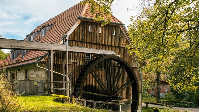 Eine historische Wassermühle mit großem Mühlrad, umgeben von Bäumen und Natur. Das Gebäude hat ein rotes Ziegeldach und Holzwände., © Stuttgart-Marketing GmbH, Sarah Schmid Eine historische Wassermühle mit großem Mühlrad, umgeben von Bäumen und Natur. Das Gebäude hat ein rotes Ziegeldach und Holzwände., © Stuttgart-Marketing GmbH, Sarah Schmid