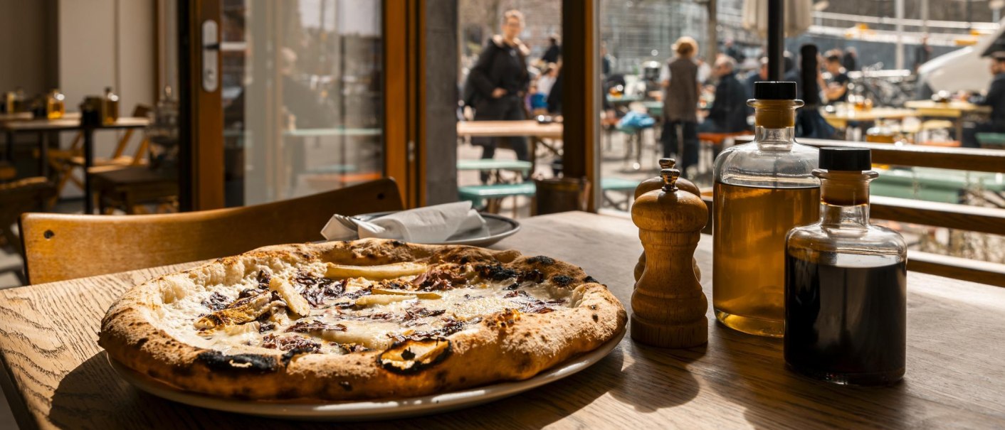 Eine Pizza auf einem Holztisch in einem Restaurant. Im Hintergrund sind Menschen auf einer Terrasse zu sehen. Auf dem Tisch stehen Ölflaschen und ein Pfefferstreuer., © SMG, Sarah Schmid