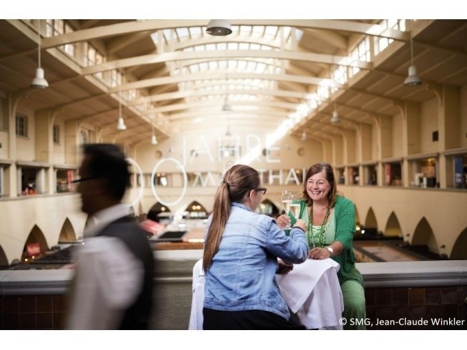 Zwei Frauen sitzen in der Stuttgarter Markthalle an einem Tisch und stoßen mit Getränken an. Ein Kellner läuft vorbei., © Stuttgart Marketing GmbH Zwei Frauen sitzen in der Stuttgarter Markthalle an einem Tisch und stoßen mit Getränken an. Ein Kellner läuft vorbei., © Stuttgart Marketing GmbH