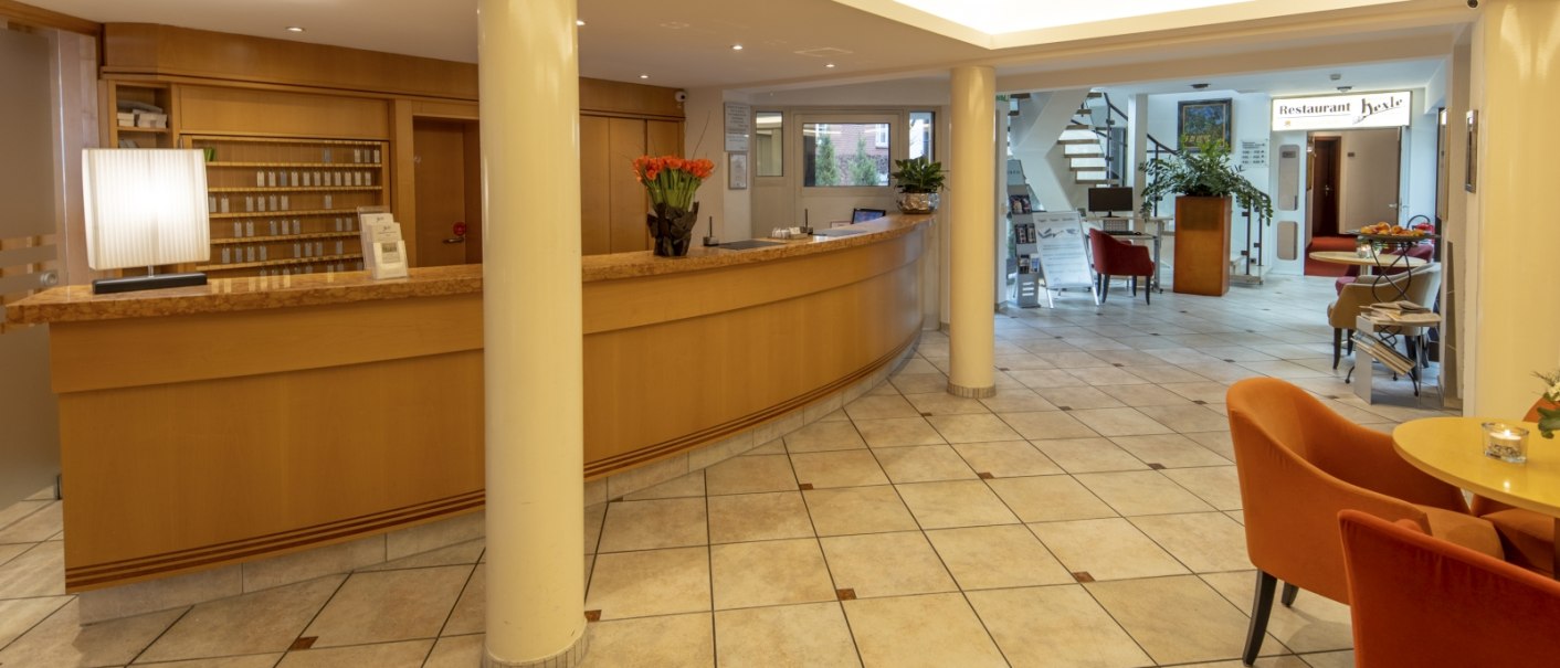 Hotel-Lobby mit geschwungener Holzrezeption, Sitzbereich mit orangefarbenen Stühlen und Hinweisschild zum Restaurant. Helle, einladende Atmosphäre., © Hotel Gloria GmbH Restaurant Möhringer Hexle