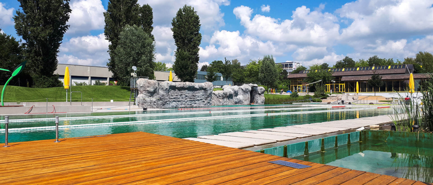 Das Naturfreibad Herrenberg zeigt einen Pool mit Holzdeck, Felsenwand und umgebender gr&uuml;ner Landschaft unter blauem Himmel mit Wolken., &copy; Stadtwerke Herrenberg