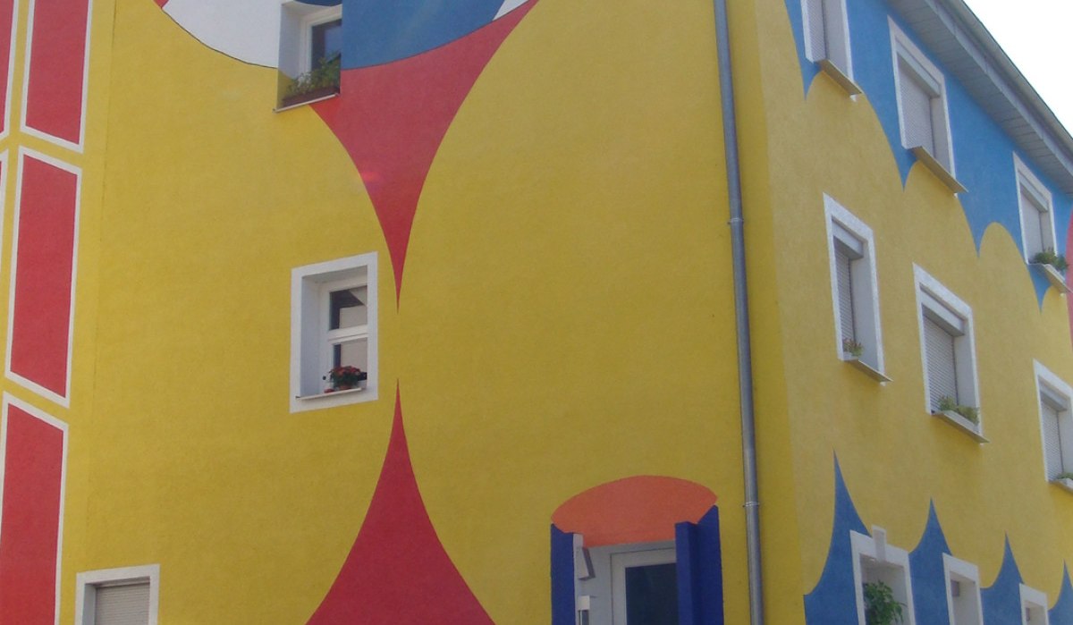 Ein Gebäude mit farbenfroher Fassade, bemalt in Rot, Gelb und Blau, zeigt geometrische Muster und ein großes Auge. Pflanzen schmücken den Eingangsbereich., © Land der 1000 Hügel - Kraichgau-Stromberg