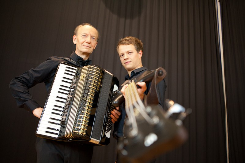 Zwei Musiker posieren mit einem Akkordeon und einem Streichinstrument vor einem dunklen Vorhang. Sie tragen schwarze Kleidung. Zwei Musiker posieren mit einem Akkordeon und einem Streichinstrument vor einem dunklen Vorhang. Sie tragen schwarze Kleidung.