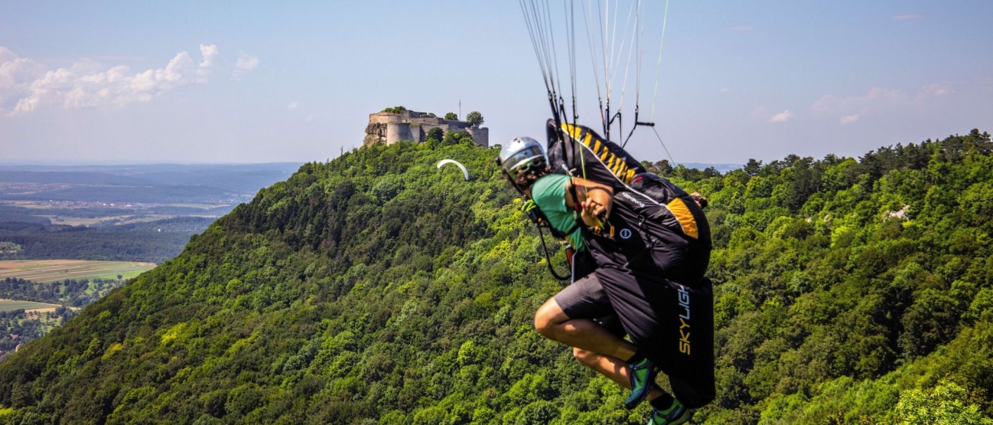 Ein Paraglider fliegt vor der Ruine Hohenneuffen über eine grüne, bewaldete Landschaft. Der Himmel ist klar und sonnig., © Stuttgart-Marketing GmbH Achim Mende
