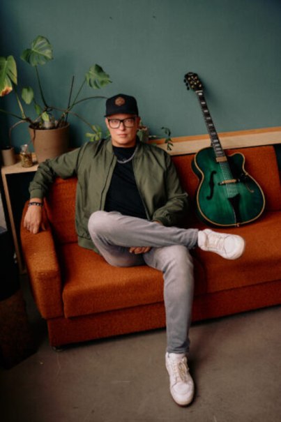 Person mit Brille und Mütze sitzt lässig auf einem orangefarbenen Sofa, eine grüne Gitarre lehnt daneben an der Wand., © Jazzclub Schwäbisch Hall e.V. Person mit Brille und Mütze sitzt lässig auf einem orangefarbenen Sofa, eine grüne Gitarre lehnt daneben an der Wand., © Jazzclub Schwäbisch Hall e.V.