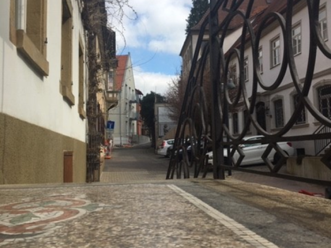 Ein kunstvolles Tor öffnet den Blick auf eine gepflasterte Straße, gesäumt von historischen Gebäuden mit roten Dächern und weißen Fassaden., © Touristik und Marketing GmbH