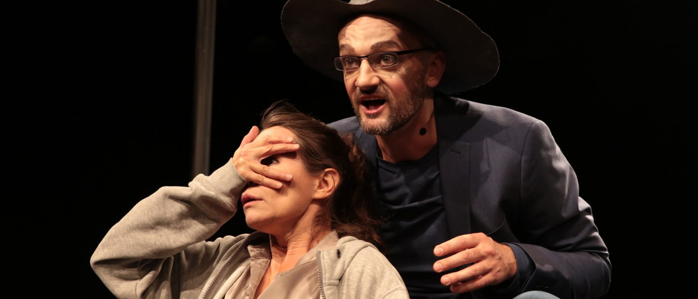 Ein Mann mit Hut und Brille spricht auf der Bühne, während er die Augen einer Frau verdeckt. Beide tragen Mikrofone., © Theater tri-bühne, Stefan Kirchknopf