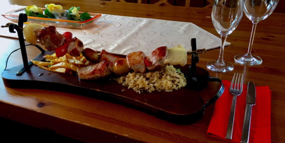 Ein Tisch mit einem Fleischspieß auf Holzbrett, Reis und Gemüse. Zwei leere Weingläser und Besteck auf roter Serviette daneben.