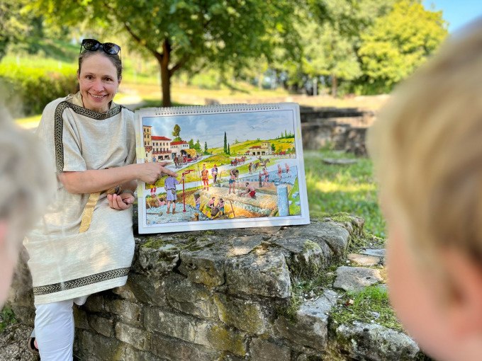 Eine Frau in römischer Kleidung zeigt Kindern ein Bild mit einer römischen Szene. Sie sitzt auf einer Steinmauer im Freien., © Stadt Nürtingen Eine Frau in römischer Kleidung zeigt Kindern ein Bild mit einer römischen Szene. Sie sitzt auf einer Steinmauer im Freien., © Stadt Nürtingen
