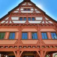 Fachwerkhaus mit rotem Holzrahmen und blauen Fenstern, umgeben von weiteren Fachwerkgebäuden, unter klarem, blauem Himmel., © SMG Achim Mende