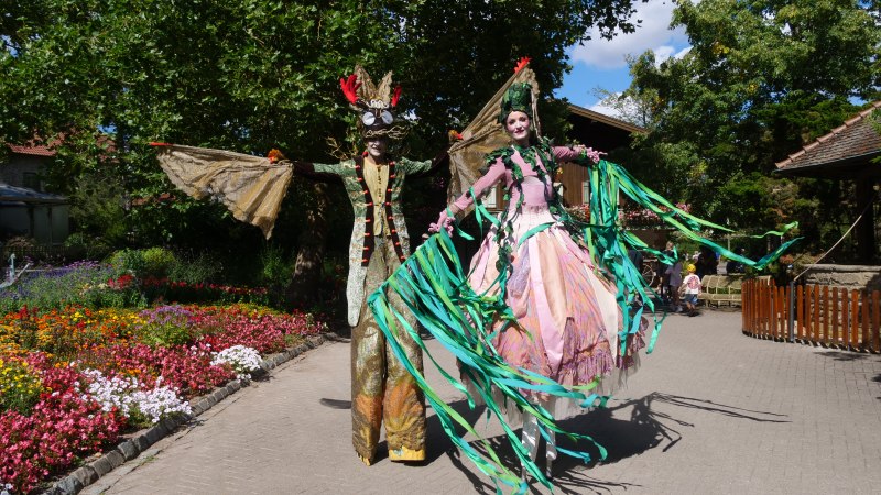 Zwei Personen auf Stelzen in fantasievollen Kostümen spazieren durch einen blühenden Garten. Bunte Blumen und Bäume umgeben den Weg., © Erlebnispark Tripsdrill Zwei Personen auf Stelzen in fantasievollen Kostümen spazieren durch einen blühenden Garten. Bunte Blumen und Bäume umgeben den Weg., © Erlebnispark Tripsdrill