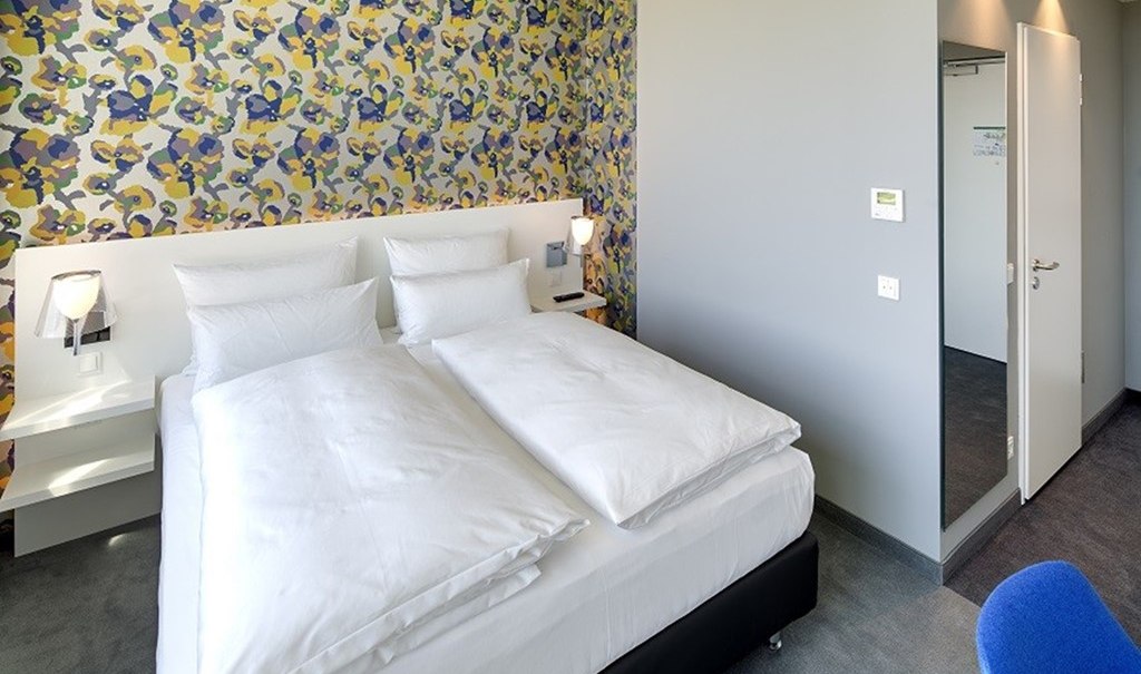 Modernes Hotelzimmer mit Doppelbett, bunter Tapete und grauen Wänden. Ein blauer Stuhl steht im Vordergrund., © Hotel am Remspark Modernes Hotelzimmer mit Doppelbett, bunter Tapete und grauen Wänden. Ein blauer Stuhl steht im Vordergrund., © Hotel am Remspark