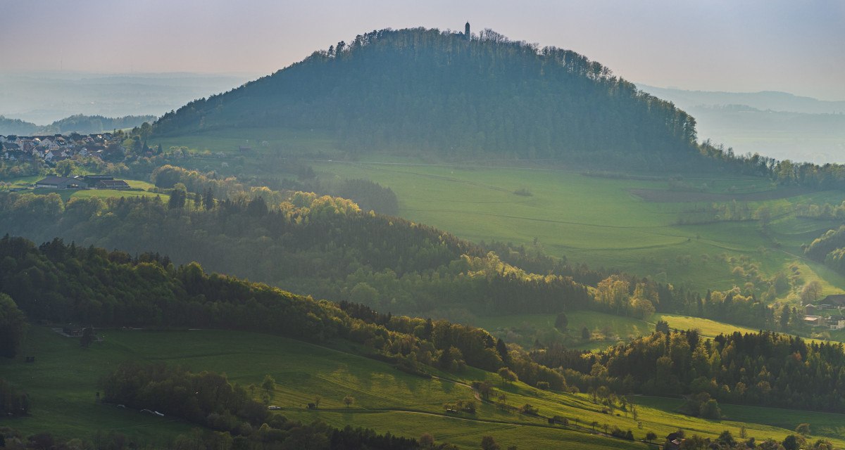 Der Rechberg erhebt sich majest&auml;tisch &uuml;ber die umliegenden gr&uuml;nen Wiesen und W&auml;lder, w&auml;hrend die Sonne sanft &uuml;ber die Landschaft scheint.