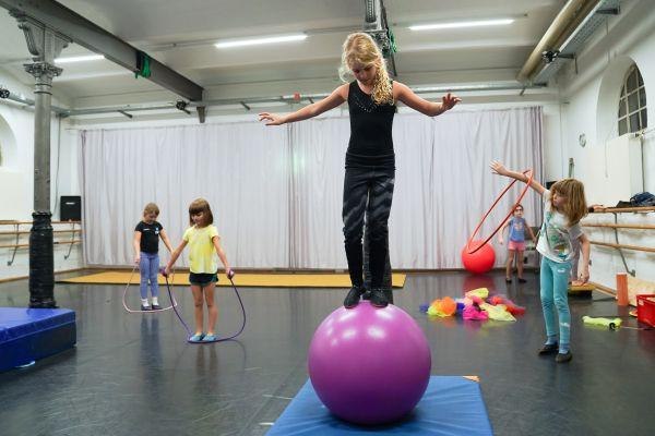 Kinder üben Zirkuskunststücke in einem Raum. Ein Kind balanciert auf einem großen Ball, während andere mit Hula-Hoop-Reifen spielen., © Kunstschule Labyrinth Kinder üben Zirkuskunststücke in einem Raum. Ein Kind balanciert auf einem großen Ball, während andere mit Hula-Hoop-Reifen spielen., © Kunstschule Labyrinth