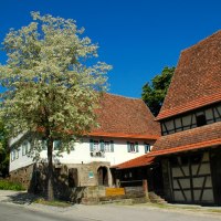 Ein traditionelles Bauernhausmuseum mit Fachwerkgebäude, blühenden Bäumen und blauem Himmel. Ein Schild zeigt Öffnungszeiten an., © Stadt Leonberg