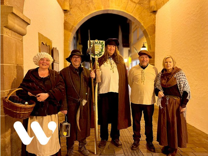 Fünf Personen in historischen Kostümen stehen unter einem beleuchteten Torbogen. Sie tragen Hüte, Umhänge und halten Laternen und Stäbe., © Stadt Winnenden