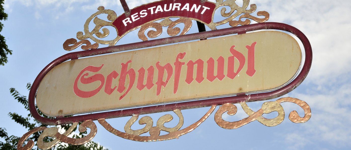 Ein dekoratives Schild mit der Aufschrift 'Schupfnudl' hängt vor einem blauen Himmel. Es gehört zu einem Restaurant., © Schupfnudl